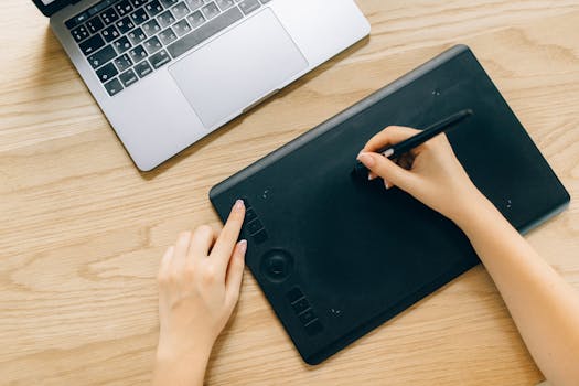 A person using a digital tablet and laptop on a wooden desk for creative work.