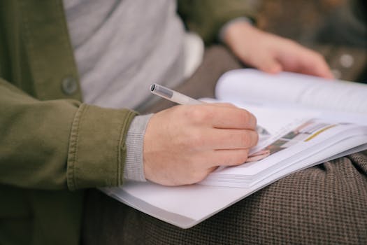 Crop unrecognizable male student in casual clothes writing in workbook while doing homework in park