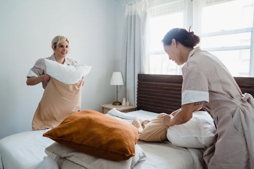 Two women in uniforms arranging bed linens in a clean and bright bedroom.